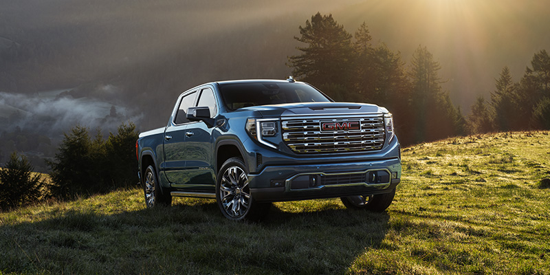 A blue 2025 GMC Sierra 1500 trekking up rugged terrain while the sun sets overhead
